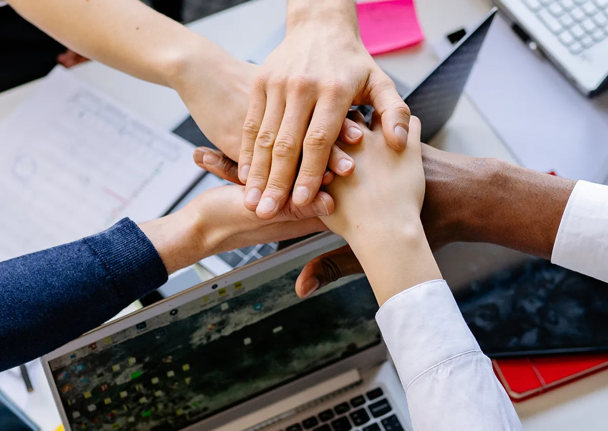 Foto von fünf Händen, die sich einvernehmlich berühren über einem Laptop. Symbolisch für ein Team. Im Blogbeitrag geht es um den Job als Community Manager